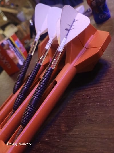 Three darts with white flights and black ribbed barrels sit in an orange holder. "PRO GRIP" is printed on the barrel of one dart. The darts are angled diagonally from the top left to the bottom right. A dark wooden surface is visible in the background. The corner of a package of snacks is visible in the top left corner. The bottom left corner shows various objects on a surface.