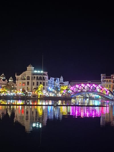 The image shows a nighttime view of a waterfront area in Phú Quốc, Vietnam, with a prominent bridge adorned with colorful lights. The lights are reflected in the dark water, creating a symmetrical and vibrant visual effect. Buildings with illuminated windows and decorative lighting line the waterfront. The scene is likely a tourist or entertainment district, given the elaborate lighting and architecture. The time of day is clearly night, and the water appears calm, suggesting fair weather. No people are visible, but the illuminated buildings suggest activity within them. The overall mood is festive and visually appealing, highlighting the beauty of the illuminated structures and their reflections. No visible text is present in the image.