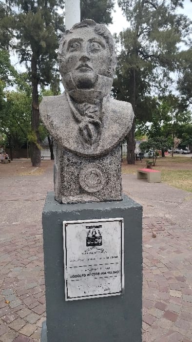A weathered bust, likely stone, depicts a man in formal attire, set atop a tall, square concrete pedestal. A plaque adorns the pedestal with text that includes "Rodolfo Hector Aiaglino". The statue is situated in a park-like environment, with trees and a brick paved ground. Benches and additional greenery are visible in the background.