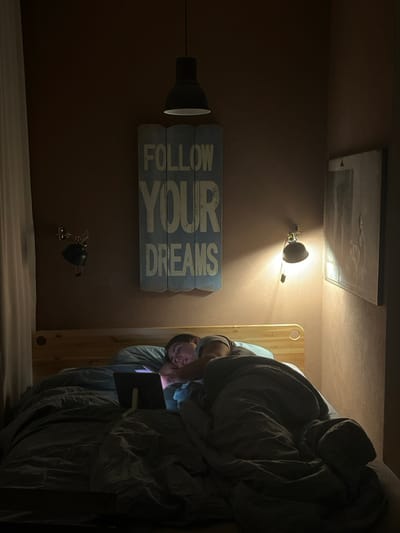A dimly lit bedroom scene in Capital City of Prague, Czechia, depicts one individual lying in bed. The person, an adult with short dark hair, is positioned on their left side with their head on a light blue pillow, partially covered by a dark grey duvet. They are wearing a light-colored t-shirt. A digital tablet, propped on a stand, rests on the bed in front of them, its illuminated screen casting light onto their face as they view content. The room features reddish-brown walls, a light wooden bed frame, and white curtains on the left. A dark pendant lamp hangs from the ceiling. Above the bed, a light blue wooden sign inscribed with "FOLLOW YOUR DREAMS" is mounted. A dark, unlit wall lamp is visible on the left, while a lit wall lamp on the right illuminates a framed picture on the adjacent wall.