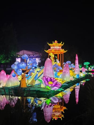 Captured at night in Puente Alto, Chile, the image presents a vibrant, illuminated art installation, possibly part of a light festival. The artwork features illuminated flowers in shades of purple, pink, yellow, and blue. A traditional orange Asian-style structure with ornate roof detailing is visible, along with a taller green structure appearing to be a central support. In the background, a building is lit with a blue and white display. The artwork is reflected in a body of water in the foreground. The overall atmosphere is dark, contrasting with the colorful lights of the installation.