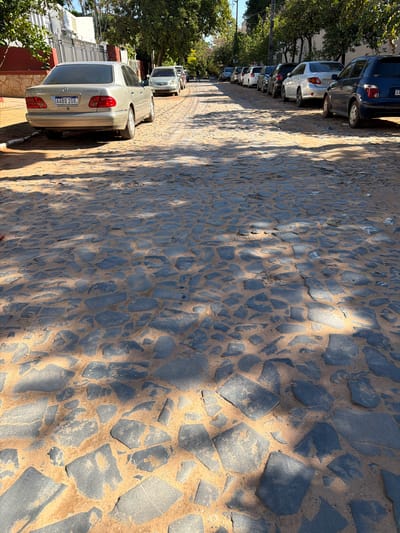 The image shows a street paved with dark cobblestones and sand. Several cars are parked along the left side of the street, with trees and buildings in the background. The sun is shining, casting shadows on the cobblestones, suggesting it is daytime. The cars are of various models and colors, and one car in the foreground has a license plate that reads "AABD 208". The scene appears to be in a residential or commercial area, possibly a street in Asuncion, Paraguay, given the location context. No people are clearly visible in the image. The overall impression is a quiet street scene on a sunny day.