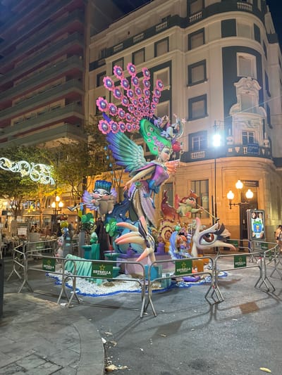 The image captures a nighttime scene in Alicante, Spain, featuring a large, elaborate festival float illuminated by streetlights and the float's own lighting. The float depicts a fantastical scene with several colorful, cartoonish figures. A prominent central figure appears to be a woman with large green wings and an elaborate headdress adorned with pink and purple flowers. Other figures include small winged cherubs, a creature with a large eye, and various whimsical characters. The float is surrounded by metal barricades and decorated with white gravel and blue accents, resembling water. In the background, modern and older buildings are visible, including a multi-story apartment building with balconies and a more ornate building with a sign for "Santander Private Banking." Streetlights and decorative lights from a nearby establishment illuminate the scene. A few people are visible in the background, including a woman walking on the right side of the frame. The overall impression is a vibrant display, likely part of a local festival or celebration.