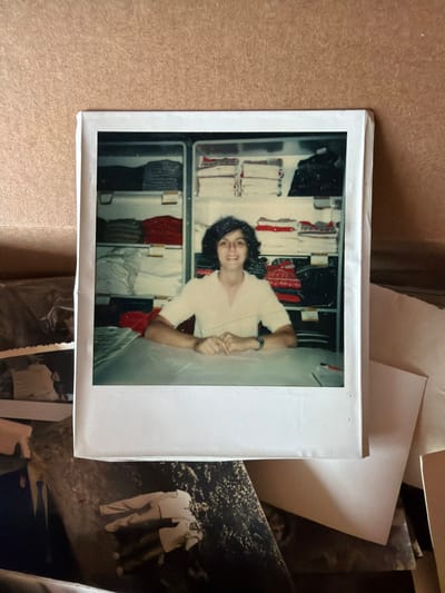 This is a physical photograph, appearing to be a Polaroid, placed on top of other photographic materials. The Polaroid features a person with dark, curly hair and a light-colored short-sleeved shirt, smiling and looking towards the camera. They are positioned in front of shelves filled with clothing, suggesting an indoor setting like a closet or a store. The person is leaning back slightly, with one arm bent and the hand resting on their hip, while the other arm is extended and bent at the elbow. The background shelves contain various items of clothing, predominantly in shades of white and red, some folded and others possibly on hangers. The overall image quality shows some signs of age and use, with a slightly grainy texture and some dust or smudges on the surface. The lighting suggests an indoor environment. To the right of the Polaroid, another photograph is partially visible, showing two people in casual attire standing outdoors, possibly in a park or garden setting. Further down, another small photo shows a person or people. The context provided indicates the location is Svilengrad, Bulgaria.