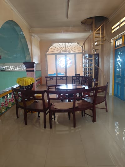 The image shows an indoor scene in a room that appears to be in Xuân Thành, Vietnam. In the center of the room, a circular dark wood table is surrounded by several dark wood chairs with curved backs. The table has an ornate, carved base and a smooth, polished surface. The chairs also feature decorative carvings.
To the left of the table, there is a decorative wall with a mural depicting a sky with clouds and stars within an arched recess. Below this arch, there is a turquoise tiled wall and a dark wood pillar with pink and white veining, topped with two objects draped in yellow fabric. Further left, there is a window with white lattice work.
To the right of the table, a spiral staircase made of light-colored metal ascends towards an opening in the ceiling. Adjacent to the spiral staircase is a tall, narrow door painted blue with decorative white accents. In the background, there is a large window with a fan-shaped decorative frame above it, revealing a circular stained-glass window. More chairs and tables are visible in the background, suggesting a dining or seating area. The floor is tiled with large, light-colored square tiles that reflect the light. The overall impression is of a well-decorated interior space with traditional elements.