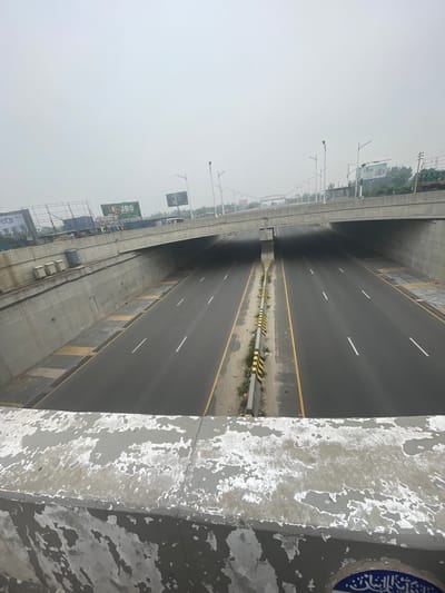 The image shows a multi-lane highway in Purbachal, Bangladesh. The highway is mostly empty, with white lane markings and yellow edge lines. A median with black and yellow striped barriers runs down the center. The roadway is under an overpass or flyover. The sky is overcast and hazy. A concrete barrier with peeling paint is in the foreground. A billboard with "JBS" is visible in the distance.