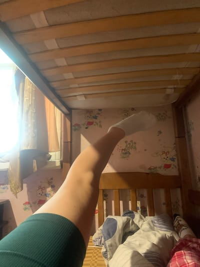 The image depicts the underside of a wooden bed frame, viewed from a low angle. A person's bare leg, wearing a green garment on the thigh, extends upwards towards the frame, with a white sock on their foot. The leg is bent at the knee. The bed frame is made of light-colored wood slats. Behind the bed, wallpaper with a pastel pink background and a repeating pattern of cartoon bunnies and flowers is visible. To the left, sheer curtains are illuminated by sunlight streaming through a window. The scene is indoors, likely a bedroom. The location of Hoholeve, Ukraine, is not discernible from the visual information present in the image.