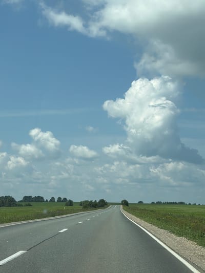 This media file captures a view from a moving vehicle on a paved road in Niva, Russia. The scene is dominated by a bright blue sky with scattered white, puffy clouds. The road stretches into the distance, curving slightly to the right, and is bordered by a grassy field on the left and a shoulder of gravel and vegetation on the right. A line of trees can be seen in the distance, bordering the field. The lighting suggests it is daytime, likely late morning or early afternoon, with fair weather. The perspective implies the photo was taken from a car window or dashboard, looking forward and slightly to the side. There are no people or specific objects visible other than the road, fields, trees, and sky. The overall impression is one of a serene, open landscape during a journey.