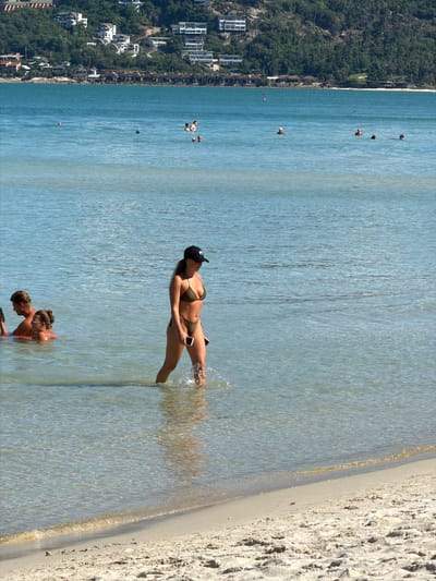 This media file depicts a sunny day at a tropical beach in Baan Chaweng, Thailand. The scene is oriented vertically, with the sea occupying the majority of the frame and a sandy beach on the right. In the water, two women are visible. One woman, wearing a dark bikini and a black cap, is standing in the shallow water, holding a smartphone and splashing water with her hand. The other woman is partially submerged, looking at her phone. Further out in the sea, a few other individuals can be seen enjoying the water. In the background, a lush green hillside is dotted with white buildings, suggesting a resort or residential area. The water is clear and turquoise, with gentle ripples on the surface, indicative of calm weather. The time of day appears to be midday, given the bright sunlight casting sharp shadows. The overall mood is one of relaxation and enjoyment of a beach vacation. No visible text is present in the image.