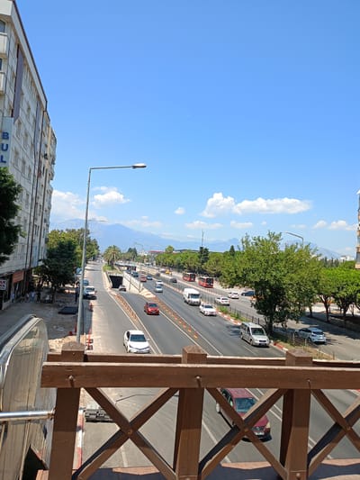 The image shows a wide, multi-lane road in Antalya, Turkey, with a clear blue sky and scattered clouds above. On the left, a modern, multi-story building with numerous windows lines the street. Trees and landscaping are visible along the sides of the road, and a range of mountains can be seen in the distance. Several vehicles, including cars and buses, are traveling on the road, and there are pedestrian walkways and streetlights. The foreground features a wooden railing, suggesting the photo was taken from an elevated viewpoint, possibly a pedestrian bridge or walkway. Construction is underway on the left side of the road, with visible building materials.