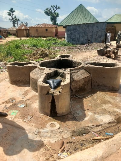 The image presents an outdoor scene in Zangon Aya, Nigeria, centered on a weathered concrete communal well structure. This primary subject consists of a central, multi-chambered basin surrounded by four cylindrical extensions, forming a cruciform or clover-leaf pattern. Water is actively spilling from an opening in the front-most cylindrical component, creating a dark, wet streak down its side and onto the surrounding ground. A dark, possibly plastic, object is partially submerged in the water within this cylinder.
The immediate ground around the well is a mix of damp soil and cracked concrete, heavily soiled with water stains, mud, and various pieces of litter including plastic fragments, wrappers, and a small white ring-shaped object embedded in the dirt. A dark shoe and its shadow are visible in the lower-left corner, indicating the perspective.
In the midground, a patch of unkempt green vegetation (weeds/grass) separates the well from the background elements. Beyond this, a structure fabricated from corrugated metal sheets is partially visible, positioned in front of a building with a distinct green, pyramidal roof. To the right of the metal structure, two adult males, dressed in long white traditional garments, are standing or walking on a dirt path. Another figure is partially visible further to the right.
The background comprises several traditional single-story houses constructed from mud or earthen materials, some featuring simple flat or slightly conical roofs. Prominent in the background are several large, mature trees with thick, gnarled trunks and sparse foliage, silhouetted against a light blue sky with scattered white clouds. Faint power lines are discernible across the sky. The scene depicts a rural or semi-urban residential area with a communal water point showing signs of heavy use and wear.