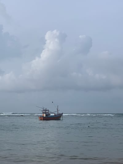 A lone fishing boat, painted orange and blue, is anchored in the calm waters off the coast of Mirissa, Sri Lanka. The sky above is overcast with large, billowy gray clouds. Gentle waves break on the distant shore. The scene is serene and tranquil, with no people visible. The fishing boat is the only subject in the frame, its weathered appearance suggesting a long history of use. The overall impression is one of quiet solitude and the vastness of the ocean.