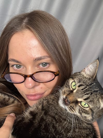 A young woman with long, brown hair and green eyes, wearing brown-rimmed glasses, is holding a tabby cat. The cat has green eyes and is nestled against the woman's shoulder. They are both looking towards the camera. The background is a grey, textured curtain. The image is a close-up selfie. The location is not specified and cannot be determined from the image.