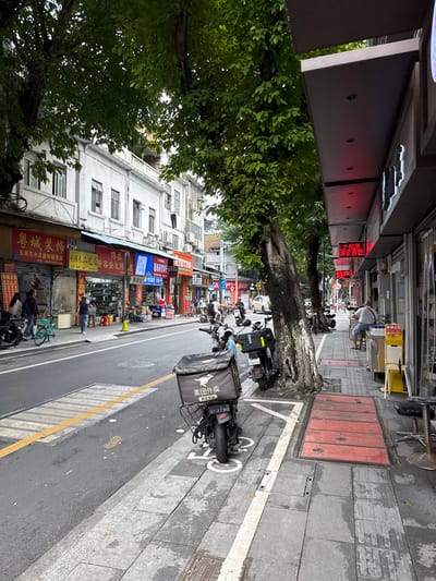 The image depicts a street scene in Liwan District, China, featuring a row of storefronts lining the right side of a moderately busy street. The architecture appears to be a mix of older, white-painted buildings with visible balconies and air conditioning units, and more modern commercial establishments with metal awnings and illuminated signs.

On the left side of the street, several trees with dense green foliage cast shadows on the sidewalk and road. The road itself is paved and marked with a yellow dividing line. Various vehicles are present, including several parked motorcycles, one of which has a large black delivery box on the back with Chinese text and the logo of Meituan. A light blue bicycle is also visible. People are seen walking on the sidewalk and crossing the street. One person is walking with a child.

The storefronts on the right side of the street display a variety of goods and services, indicated by signage in Chinese. Some shops appear to be clothing stores, while others might be food establishments or other retail businesses. There are tables and chairs set up outside one of the establishments, suggesting outdoor seating. A person is seated on a stool outside a shop. The sidewalk is paved with grey tiles, and there are metal covers on the ground, possibly for utility access. The overall atmosphere is that of a typical urban commercial street.