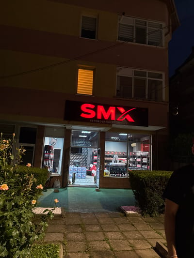 A two-story building in Targovishte, Bulgaria, is depicted at night, illuminated by artificial light. The building's facade features horizontal bands of light yellow and reddish-brown. Multiple windows are present on both floors; one upper-story window displays a warm orange glow emanating from behind closed blinds.
The ground floor hosts a commercial establishment, prominently identified by a red illuminated sign that reads "SMX" with a stylized 'X', and beneath it, "АВТОМОБИЛНА КОЗМЕТИКА" (Automobile Cosmetics). The shop's interior is visible through large glass windows and an open central doorway. Inside, extensive shelving units are stocked with various products, including items in red packaging and stacks of blue square containers on the floor. A white stepladder is visible. The interior also features a wall with a geometric pattern and a mounted flat-screen monitor. A cluster of red and white balloons is situated near the back of the shop's right side.
The exterior foreground includes a paved walkway composed of square concrete tiles, with weeds growing in the interstices. A patch of light green flooring or ground covering extends from the shop's entrance. To the left, a dense rose bush with several pale orange/peach roses in bloom occupies a garden bed, bordered by white decorative stones. A neatly trimmed green hedge lines the walkway on the right side. A small rectangular bed with pink/purple decorative stones is positioned in front of the hedge. Partially visible on the far right edge of the frame is the right shoulder and arm of a person, wearing a dark t-shirt and a silver bracelet on the wrist.