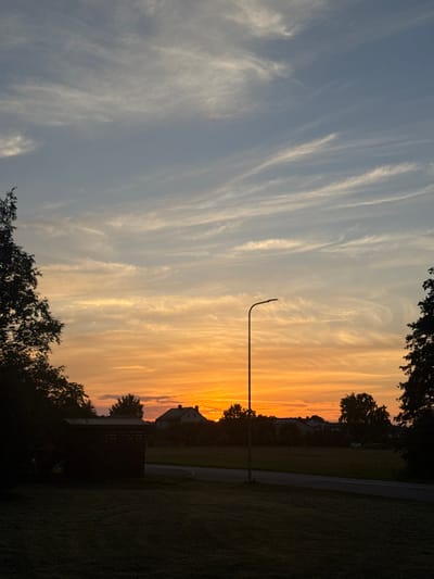 This is a vertical, eye-level shot of a sunset over a rural area in Carnikava, Latvia. The sky transitions from a pale blue at the top to vibrant orange and yellow hues near the horizon, with wispy white clouds scattered throughout. A modern-style streetlamp stands silhouetted against the colorful sky on the right side of the frame, its arm extending to the right. In the foreground, a dark, manicured lawn leads to a paved road that curves gently to the right. To the left of the road is a dark, rectangular structure, possibly a shed or small outbuilding, also silhouetted. In the middle ground, the silhouettes of trees and houses are visible against the sunset. A white car is parked in the distance on the right side of the road, near a cluster of houses. The overall scene depicts a tranquil evening in a suburban or rural Latvian setting.