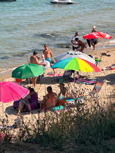 The image depicts a sunny day at a beach in Ravda, Bulgaria. Several people are enjoying their time near the water. In the foreground, a person lies on a lounge chair under a pink umbrella. To their left, a green umbrella shades another person, who appears to be talking to someone holding a unicorn-shaped inflatable.
Further back, several individuals are seen relaxing on the sand. One man in blue shorts is standing near the water, looking towards the sea. Another person is partially visible behind a beach umbrella. A red umbrella with "Coca-Cola" text is open, and a person is standing beneath it. The beach is sandy, and the water is calm with gentle waves.
There are several beach umbrellas of various colors, including red, yellow, green, and blue, providing shade to the beachgoers. Towels and bags are scattered on the sand. The scene suggests a leisurely day at the beach, with people sunbathing, relaxing, and interacting. The time of day appears to be daytime, indicated by the strong sunlight and shadows. The presence of beach umbrellas and people in swimwear confirms the beach setting.