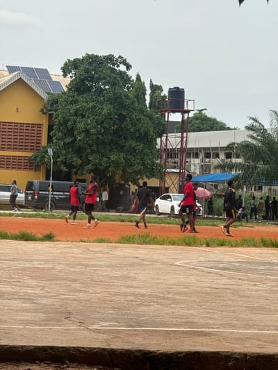 In Amansee, Nigeria, the image depicts a group of young men on an outdoor sports court, likely a basketball or volleyball court given the markings visible on the concrete surface. The court has a red dirt perimeter with green grass growing along the edge. The men are dressed in athletic wear, with some wearing red vests over black shorts and others in black shirts and shorts. Some are engaged in what appears to be a practice or game, with one individual dribbling a ball and others in motion.
In the background, a yellow building with a red brick facade and solar panels on its roof is visible. Adjacent to this building is a tall water tank supported by a red metal structure. Further in the background, construction is underway on another building, with scaffolding and white sheeting. Several vehicles, including a black van with text on its side and a white car, are parked near the yellow building. A group of people are standing and observing the activity on the court, some under a pink umbrella. The sky is overcast. The overall scene suggests a sports facility at an educational institution or a community center in Amansee.