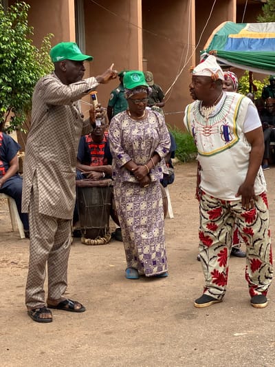 This image captures an outdoor community event in Shaba, Nigeria, likely commemorating World Environment Day 2021. In the foreground, a man wearing a light brown checkered suit and a green baseball cap is actively speaking into a microphone, gesturing with his right hand. His cap clearly displays "WORLD ENVIRONMENT DAY" and "JUNE 5 2021". Standing next to him is a woman dressed in a purple and white patterned traditional outfit, also wearing a green baseball cap with similar text, indicating her participation in the event.
Further back and to the right, another man stands prominently. He is adorned in a white tunic with intricate colorful embroidery, paired with patterned trousers featuring red and black floral motifs. He wears a distinctive white headwrap with colorful tassels and a necklace made of shells or beads, suggesting traditional or ceremonial attire. His gaze is directed downwards. Behind the speaker and the woman, a man is seated next to a traditional African drum, suggesting musical accompaniment or cultural performance.
The scene is set in an open outdoor area with a light brown ground, possibly paved or earthen, and a plain light brown building or wall in the background. Green foliage from a bush is visible in the lower-left corner. Colorful banners or fabrics (green, blue, yellow, white) hang vertically in the upper-left background. The natural lighting indicates it is daytime, under clear weather conditions. The overall atmosphere suggests a community gathering or cultural celebration.