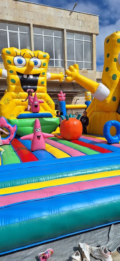 This image displays an outdoor scene in the city of Letnitsa, Bulgaria, centered on a large, multi-level inflatable play structure. The inflatable is predominantly yellow and designed as a Spongebob Squarepants bounce house, featuring two prominent Spongebob figures. Several pink inflatable Patrick Star figures are distributed across the structure's colorful surface, which includes sections striped with blue, green, yellow, pink, red, and light blue.
Multiple children are visible on the inflatable: one girl in a pink outfit stands on an elevated platform adjacent to a pink Patrick figure, while other children interact with various parts of the structure, including an orange spherical inflatable and a blue inflatable ring. A large, light-colored building constructed of stone or concrete forms the backdrop, presenting multiple large, white-framed windows that contain internal string lights. The sky above is partly cloudy blue. In the immediate foreground, several pairs of footwear, including pink child's sneakers and white adult athletic shoes, are arranged on a grey tarp next to the inflatable.