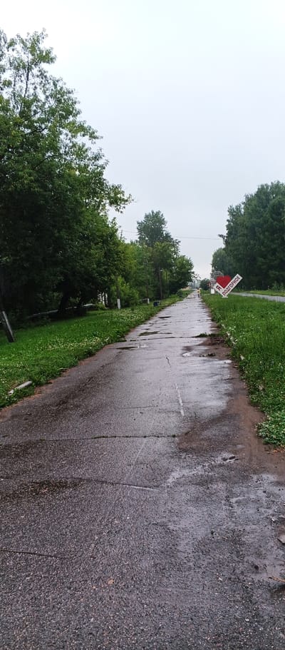The image depicts a wet, paved road in the city of Podgornoye, Russia. The road is lined with lush green grass and trees on both sides, suggesting a natural or park-like setting within the city. The sky is overcast and appears to be a bright, hazy white. On the right side of the road, a sign with a red heart shape and the words "Я ПОДГОРНОЕ" (I love Podgornoye) is visible, indicating a local landmark or tourist attraction. A person, likely a pedestrian, is walking in the distance on the road. The overall scene conveys a somber or tranquil atmosphere due to the weather and the muted colors.