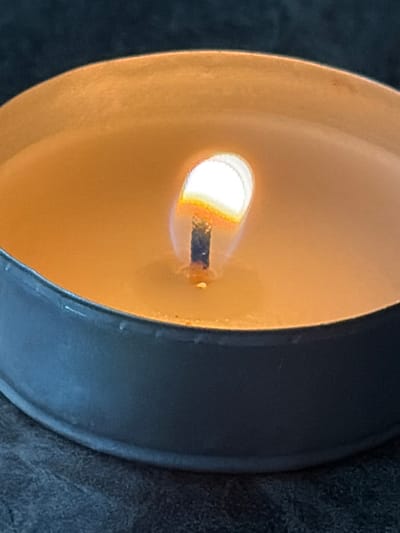 A close-up, high-angle view depicts a lit tea light candle, situated in Saint-Étienne, France. The candle rests within a dark gray or black circular metal holder, containing a shallow, molten pool of pale yellow wax. A charred wick extends vertically from the wax, supporting a bright, teardrop-shaped flame. The flame's core is intensely white, transitioning to yellow, orange, and faint blue at its base and edges, indicating active combustion. The interior surfaces of the metal holder and the surrounding melted wax exhibit a warm, reflective glow from the flame. The background is dark, undifferentiated, and out of focus, suggesting an indoor environment with low ambient light.