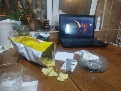 The image depicts a close-up, overhead view of a wooden table surface. In the foreground, a glass filled with a dark, frothy beverage sits to the left. A partially opened bag of potato chips is spilling onto the table, with several chips scattered around it. To the right of the chips, a pack of cigarettes with a lighter placed on top is visible. Further to the right, an ashtray containing crumpled tissues and ash is present. In the background, a laptop is open, displaying an image on its screen. Behind the laptop, various objects are situated on a shelf, including plants in pots, jars, and a bottle with liquid. The environment appears to be indoors, possibly a living space or a room. The overall impression is one of a casual or relaxed setting.