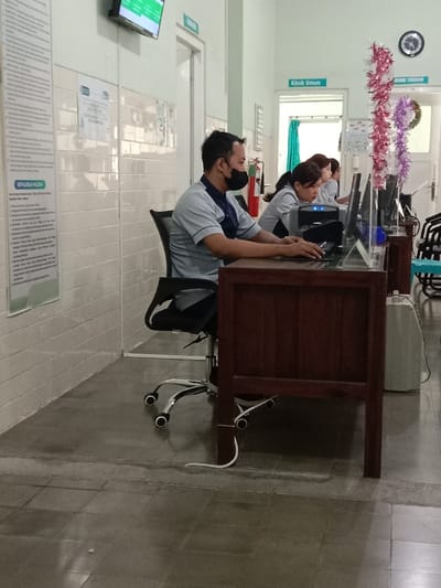 The image depicts an indoor administrative or reception area, likely within a medical facility in Madiun, Indonesia, although the specific address or name of the facility is not discernible.

The scene features three individuals working at computer workstations. In the foreground on the left, a male individual is seated in a black office chair with a chrome base. He wears a light grey polo shirt with a dark blue collar and a black face mask, and is actively using a computer mouse and keyboard on a dark wooden desk. Behind and to the right of him, two female individuals are partially visible, similarly dressed in light grey uniforms and engaged with their own computer monitors, keyboards, and mice at adjacent dark wooden desks. A clear acrylic partition separates the foreground desk from the others.

The environment includes light-colored walls, with a lower section covered in white horizontal tiles. Several signs in Indonesian are mounted on the walls, including "KLINIK UMUM" (General Clinic) over a doorway in the background, "RUANG TINDAKAN" (Procedure Room) over another, and text panels on the left wall detailing "PENULISAN RESEP" (Prescription Writing) and "KEWAJIBAN PASIEN" (Patient Obligations). A digital screen displaying green text is mounted high on the left wall. A red fire extinguisher is visible on the floor against the wall between the first two workstations. Decorative pink, red, purple, and white tinsel streamers hang vertically between workstations towards the right side of the frame, alongside a green wreath and a wall clock. The floor is covered with large, grey-toned tiles. A white electrical cable extends from beneath the foreground desk and is secured to a desk leg before resting on the floor. A white, possibly portable, air conditioning unit is visible on the floor to the far right. All individuals appear focused on their computer tasks.