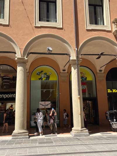 The image shows a street scene in Bologna, Italy, featuring a series of arched storefronts. The buildings are peach-colored with white trim around the windows. There is a LEGO store visible, indicated by the LEGO logo and displays in the windows. People are walking in front of the stores, some looking into the windows. A "goldenpoint" store is also visible. The architecture includes arched openings supported by pillars.