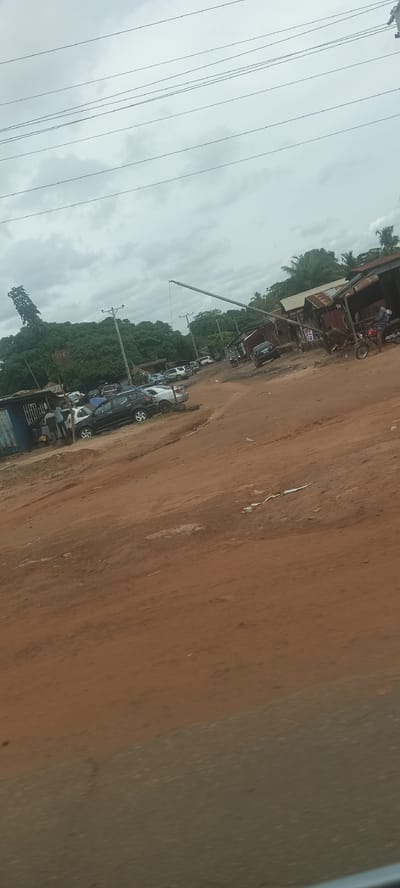 The image depicts an unpaved road intersection in Markurdi, Nigeria. The road is primarily red dirt. Several vehicles are parked on the left side of the road. A group of people stand near a blue corrugated metal structure. In the background, a large tree is visible, along with a crane-like structure extending into the sky. On the right side, there are makeshift stalls with rusty metal roofs. A person sits on a motorcycle near one of the stalls. Electrical wires are strung across the sky. The sky is overcast and gray.
