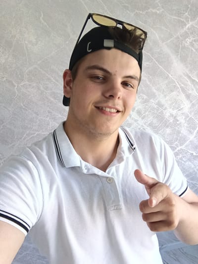 A young man stands indoors, facing the camera. He wears a white polo shirt with black stripes on the collar and sleeves, and a black baseball cap with sunglasses perched on top. He is smiling and pointing directly at the viewer with his right hand. The background is a gray wall with a marble-like pattern. The location is Andreyevo, Russia.