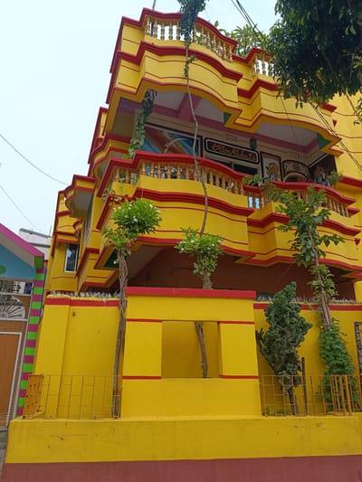The image shows a vibrantly colored, multi-story building in Benapole, Bangladesh. The building is predominantly yellow with red trim and decorative elements. The architectural style is ornate, featuring multiple levels with balconies and railings. Several small trees and some foliage are visible around the building. In the background, there is a cloudy sky and glimpses of other structures. The scene appears to be a residential or commercial building in an urban or semi-urban environment.