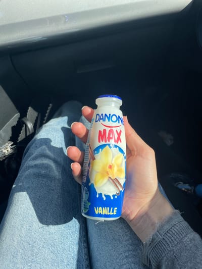 The image depicts a close-up view of a person's left hand holding a white plastic bottle of Danone MAX Vanille yogurt drink. The hand exhibits manicured fingernails with white tips. The person is wearing light blue denim jeans and a grey long-sleeved top with a visibly frayed cuff on the right wrist. The Danone bottle has a blue screw cap and a label featuring a yellow vanilla flower and a vanilla bean against a white liquid splash on a blue background, with "DANONE" in blue, "MAX" in red, and "VANILLE" in yellow text. The setting appears to be inside a vehicle, indicated by dark interior surfaces and a lighter grey panel visible in the upper left. Strong sunlight casts clear shadows within the vehicle's interior. A dark bag or purse with a silver buckle is partially visible to the left of the person's leg. This scene is located in Skhirat, Morocco.