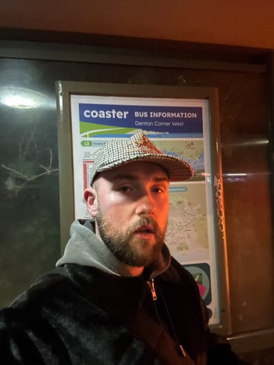 A man stands in front of a bus information sign located in Newhaven, United Kingdom. The sign reads "coaster BUS INFORMATION Denton Corner West". The man is wearing a black and white checkered hat, a grey hooded sweatshirt, and a dark jacket. The photograph appears to be taken at night. The bus information sign includes a map.