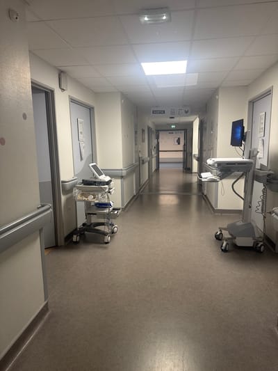 The image depicts a brightly lit hospital hallway. Medical equipment on wheels is positioned along the right side of the corridor. The walls are painted white, and doors are visible along the left side, some with room numbers or signs on them. A sign with the word "SORTIE" and directional arrows is visible near the end of the hallway. The flooring is a patterned grey color. The scene's location is inside a hospital in the city of Tourcoing, France. There are no people visible in the image.