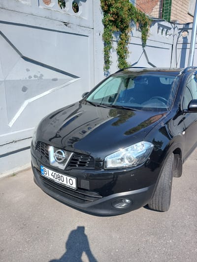 A black Nissan Qashqai is parked in front of a light grey concrete wall with a decorative pattern and some overgrown green ivy. The car has a license plate that reads "BI 4080 IO". The license plate also contains text that appears to be "UA" and a phone number "(0362) 460 999". The front of the car, including the grille and headlights, is clearly visible. The ground is asphalt, and a shadow of a person holding a phone, presumably the photographer, is cast on the ground in front of the car. The scene appears to be a street or parking area in Kremenchuk, Ukraine.
