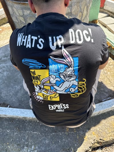 A person is wearing a black T-shirt with a Bugs Bunny design and the phrase "WHAT'S UP DOC!" in white lettering. The design includes Bugs Bunny images in blue and yellow squares. The person is standing outdoors on a concrete surface with textured stone or mosaic-like sections. The location is in the city of Probuda, Bulgaria.