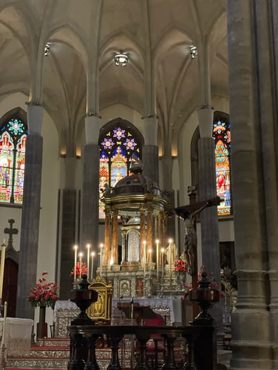 The image displays the grand interior of a church or cathedral in San Cristóbal De La Laguna, Spain. The scene is viewed from an aisle, looking towards the main altar.
**Subjects:** The dominant architectural features include tall, fluted stone columns with decorative capitals, which support elaborate vaulted ceilings in a light, neutral color. Multiple stained glass windows are visible, including round rose windows with intricate patterns and rectangular ones depicting religious figures and scenes. At the focal point is a highly ornate main altar or retable, featuring rich gold and silver elements. Above the altar stands a prominent crucifix with the figure of Christ, draped with small red and white pennants. A gold-colored structure, possibly a tabernacle or monstrance, is central to the altar. Another religious figure (possibly a statue) is partially visible to the left.
**Activity or event taking place:** No people are visible, and no specific activity or event is ongoing, presenting a quiet and contemplative scene within the sacred space.
**Notable details:** The interior is dimly lit, with light primarily emanating from the vibrant stained glass windows, suggesting it is daytime. Lit candles on the altar contribute to the ambiance, and arrangements of red flowers adorn the altar and a nearby pedestal, adding color to the solemn setting. The inscription "INRI" is clearly visible above the figure of Christ on the crucifix. The grandeur of the architecture and the religious artifacts evoke a sense of reverence and spiritual solemnity.