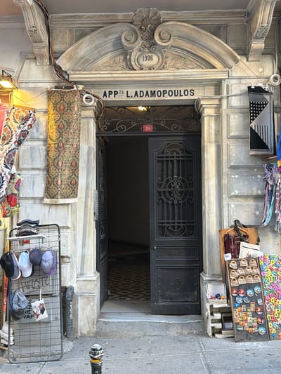 An exterior shot captures a grand, weathered entrance to an apartment building in Beyoğlu, Turkey. The facade is adorned with neoclassical architectural details, including ornate carvings and a stone archway above the doorway. The year "1906" is prominently displayed within a carved medallion, hinting at the building's age. The name "APPS L. ADAMOPOULOS" is inscribed above the entrance.

The dark, wrought-iron double doors of the entrance are partially open, revealing a dimly lit interior with a patterned floor. A red plaque with the number "24" is affixed to the door. To the left of the entrance, several colorful textiles, possibly rugs or tapestries, are hanging. A rack filled with stacked caps and hanging baseball caps is also present, suggesting a shop or market stall.

To the right of the entrance, a collection of items is displayed on the sidewalk. This includes framed items with small pictures and text, likely souvenirs, as well as a large board covered with colorful letter magnets. Various bags and other merchandise are also visible. A small, decorative bollard stands on the sidewalk in the foreground. The overall scene depicts a bustling urban environment with elements of historical architecture and commercial activity.