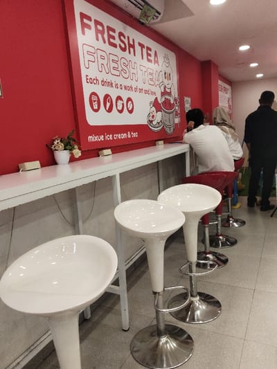 The image shows the interior of a tea shop in Bekasi Regency, Indonesia, located at coordinates -6.338 latitude and 107.166 longitude. The shop has a red wall with a large sign advertising "Fresh Tea Fresh Tea" and promoting the idea that "Each drink is a work of art and love."  The sign also features a cartoon image of a person wearing a red hat, holding a cup of tea and sitting on a stool. The sign has icons for a cup, a straw, a cow, and a coffee bean.  A white counter sits in front of the wall and has a white tabletop and white legs. The counter is topped with a row of white bar stools with shiny silver bases. There are four stools, two facing the wall and two facing the center. The stools are empty and have a white, smooth, ceramic top.  A red bar stool is partially visible in the background.  The floor is tiled in light gray and has a gray grout. 
Three people are visible in the background: a woman wearing a gray shirt is seated on the red bar stool, a man is seated on a white stool, and a man is standing near the far wall wearing a black shirt and dark pants. All three people are facing the wall. There is an abundance of light in the room, giving the interior a bright, clean feel. The shop appears to be in the midst of a business day, with patrons present and likely ready to order tea and enjoy the atmosphere.