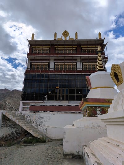 The image shows a view of the Phyang Monastery in Phyang, India, a Buddhist monastery located in the Indus Valley. The monastery is situated on a hillside and is characterized by its whitewashed walls, red and gold accents, and intricate architectural details. The main building of the monastery is a multi-story structure with numerous windows and decorative elements on its roof. In the foreground, there are several stupas, which are Buddhist reliquary monuments. One prominent stupa in the foreground is white with a striped conical top and colorful decorative bands. Adjacent to the stupa, there are stairs leading up to the monastery. The sky is cloudy with patches of blue, suggesting it may be a partly sunny day. The surrounding landscape appears to be mountainous and arid, typical of the region.