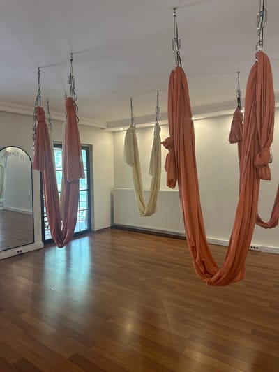 The image shows the interior of a yoga studio. Aerial yoga hammocks in peach and off-white colors are suspended from the ceiling. The floor is wooden. A window and a mirror are visible in the background. The location is likely in Esenyurt, Turkey, given the user's location. No people are visible in the image. The scene appears to be set up for aerial yoga practice.
