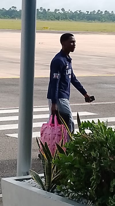 A young Black man walks across an airport tarmac, holding a pink tote bag in his left hand and a dark-colored smartphone in his right. He is wearing a navy blue long-sleeved shirt with graphics on the sleeves, including what appears to be a Union Jack flag patch on the left sleeve and "LONDON" text on the right sleeve. He also wears light blue pants. The man is walking on a striped pedestrian crossing. In the background, there is an airport runway, a line of trees, and a cloudy sky, suggesting it is daytime. The man appears focused as he walks. The scene takes place at an airport in Orerokpe, Nigeria.