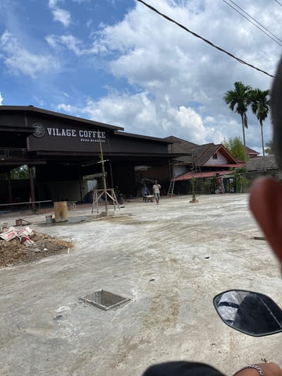 The image depicts an outdoor scene in front of a building with a sign reading "VILLAGE COFFEE Feat Acoustic". The building appears to be under construction or renovation, with scaffolding and construction materials visible. Two men are present, one pushing a wheelbarrow and another walking. A motorcycle is parked to the right of the building. The ground is paved with concrete, and there is a square manhole cover in the foreground. Palm trees are visible in the background, suggesting a tropical or subtropical climate. The sky is partly cloudy. The presence of the "VILLAGE COFFEE" sign indicates a commercial establishment. The overall environment suggests a business operating in a developing area.