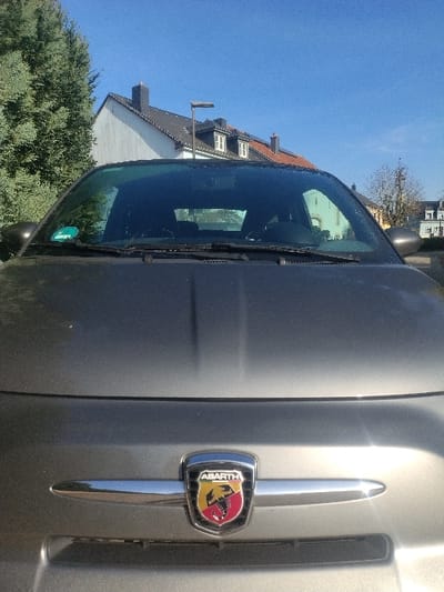 The scene depicts the front of a grey Abarth automobile, identifiable by its scorpion emblem on the grille, situated outdoors in Crauthem, Luxembourg, under a clear blue sky. The vehicle's hood displays two subtle longitudinal raised lines. The windshield, equipped with wiper blades, has a green sticker affixed to its lower left side. To the immediate left of the vehicle is a large, dark green coniferous tree. In the background, multiple residential-style buildings are visible, including a white structure with a dark grey tiled roof and chimney, and another building with an orange-red tiled roof featuring dormer windows. A dark grey street light pole stands between these background structures. Further right, distant buildings and a bare deciduous tree are partially visible. The setting is illuminated by bright daylight, casting reflections on the car's metallic surfaces. No individuals are present within the frame.