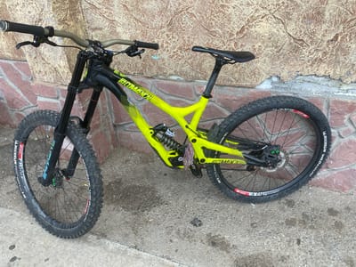 A bright yellow and black Commencal downhill mountain bike is parked outdoors, leaning against a textured wall. The bicycle features a full suspension setup with a black dual crown fork at the front and a coil shock near the bottom bracket. Its frame is predominantly vivid yellow/lime green with black graphics and accents.
Both front and rear tires are Michelin Wild Enduro, with the front tire specified as "GUMXT 2.40" and the rear as "GUMXD TL 2.40." The rims are DT Swiss, with the front identified as "FR570" and the rear as "EX471." The bike is equipped with disc brakes, and the front caliper has noticeable green bolts or accents. The handlebars are wide and appear to be an earthy green/bronze color, fitted with black grips. A SRAM logo is visible on the right shifter. The bike has purple/pink flat pedals. The black saddle is mounted on a black seat post.
The background consists of a textured wall with varied shades of brown, beige, and possibly some salt efflorescence or peeling paint. The ground is grey concrete, showing some stains and debris. The image appears to be taken during the daytime under clear and dry weather conditions. The bike is stationary, and no activity is occurring. The location is Troyan, Bulgaria.