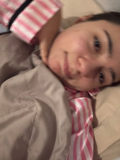 A blurry, close-up selfie captures a young woman with dark hair and fair skin. She lies in bed, covered by a light brown comforter. Her pajamas, striped in pink and white, are visible at the edges of the frame. The background is indistinct, hinting at a bedroom setting with a pillow. The overall image has a relaxed, informal feel, suggesting a candid moment.