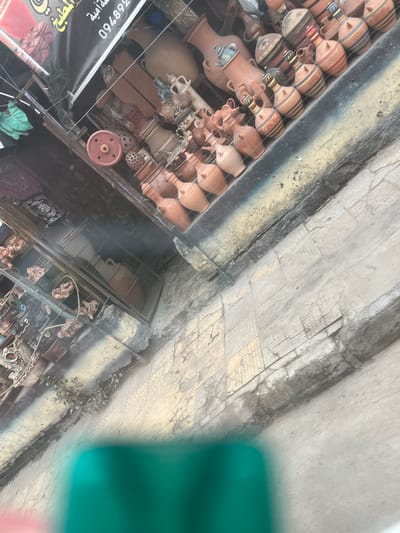 An array of earthen vessels, including vases and pitchers, are arranged on a tiered display within a shop. Their forms range from small, simple shapes to larger, more ornate designs. The shop's awning is visible, with some signage. The adjacent sidewalk is cracked and uneven, leading away from the display. Other goods, possibly textiles, are partially visible below the earthen pots.