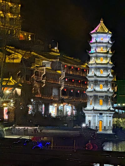 This nocturnal image displays a segment of the ancient town of Fenghuang, China, illuminated along a waterway. On the right, a multi-tiered pagoda, exhibiting traditional Chinese architectural characteristics, is brightly lit with a gradient of colors including pink, yellow, blue, and orange. Its lower sections are stone-colored with several arched windows emitting warm yellow light.

To the left of the pagoda, a series of multi-story traditional buildings with dark tiled roofs and ornate wooden balconies ascend a hillside. These structures are extensively lit with warm yellow artificial light emanating from within, revealing what appear to be retail spaces on their lower levels. Numerous red lanterns hang from the balconies of these buildings.

In the foreground, a dark body of water, presumably a river or canal, reflects the urban illumination. Several elongated, flat-bottomed vessels or rafts are present on the water, some decorated with prominent blue and red accent lights. A single individual is discernible near the base of the pagoda on a waterfront platform, holding a white illuminated umbrella. A streetlamp with a bright white light is visible above the buildings on the upper right. The night sky is uniformly dark, providing a backdrop for the illuminated scene. Bare tree branches are partially visible on the far left.