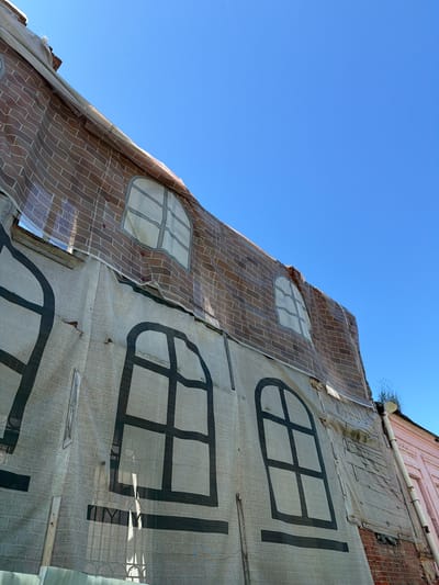 This image shows a close-up, low-angle view of a building facade covered in tarpaulin. The tarpaulin is printed with a trompe-l'œil design simulating a brick wall with several windows. The printed windows are drawn in a simplified, dark outline style, with some appearing to have panes. One section of the tarpaulin depicts a brick pattern with a light grey background and darker grey bricks arranged in a staggered pattern. Another section is a lighter beige color with a grid-like pattern and larger, dark outlined window shapes.
The building itself is partially visible behind the tarpaulin on the right side of the image. It appears to be constructed of brick and has some architectural details like a white pipe running horizontally along the facade. There are small gaps and tears in the tarpaulin, revealing the actual building material underneath, and some wooden planks are visible, possibly used for support or repair. Small, sparse branches with green leaves are growing from the top edge of the building, suggesting it's a warm or temperate season.
The scene is illuminated by bright sunlight, casting shadows and creating highlights, indicating it is daytime. The sky is a clear, vibrant blue, suggesting fair weather. The angle of the sun and the shadows cast on the tarpaulin suggest it is likely midday. The overall impression is that of a building undergoing renovation or being temporarily covered for protection, with the printed facade creating an illusion of a completed structure. The location context of Mahilyow, Belarus, is noted. No visible text is present in the image.